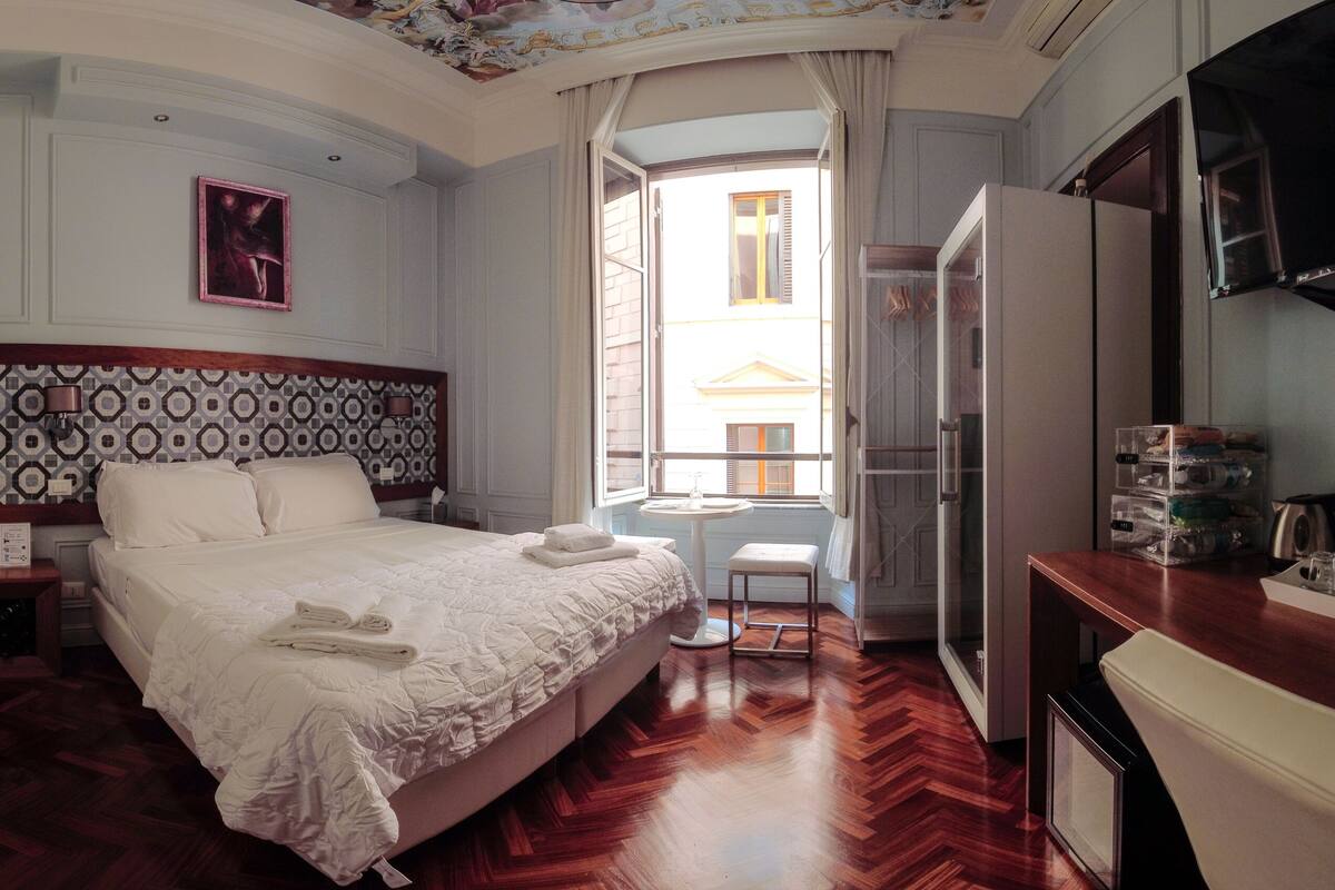 A hotel room with a large bed, a desk, a chair, a small table, a TV, and a view of the outside.