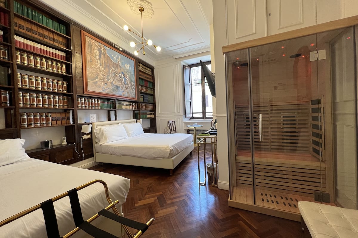 A hotel room with a large bookshelf, a bed, a desk, and a glass shower enclosure.