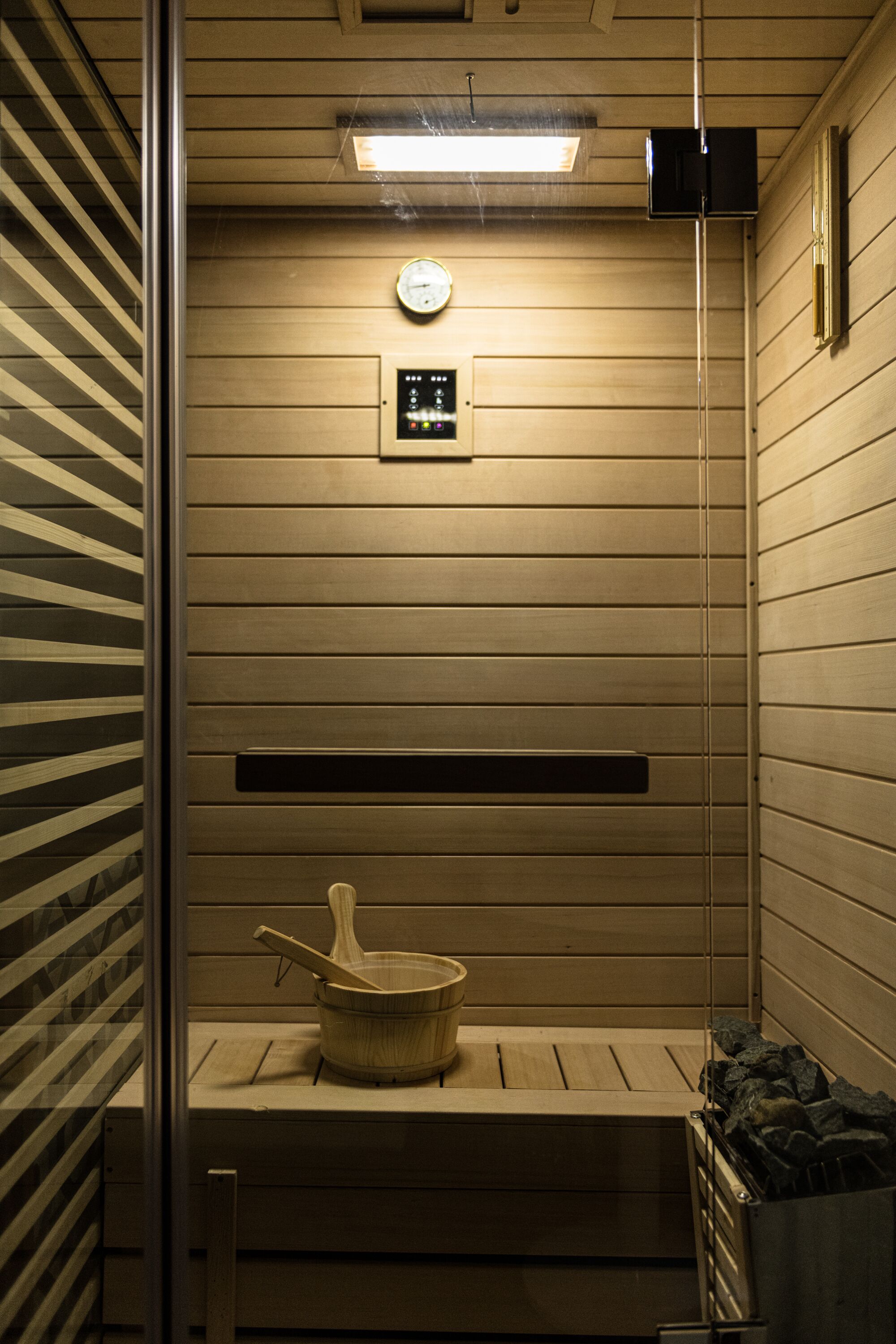 A sauna room with wooden walls, a ceiling light, a clock, and a control panel.