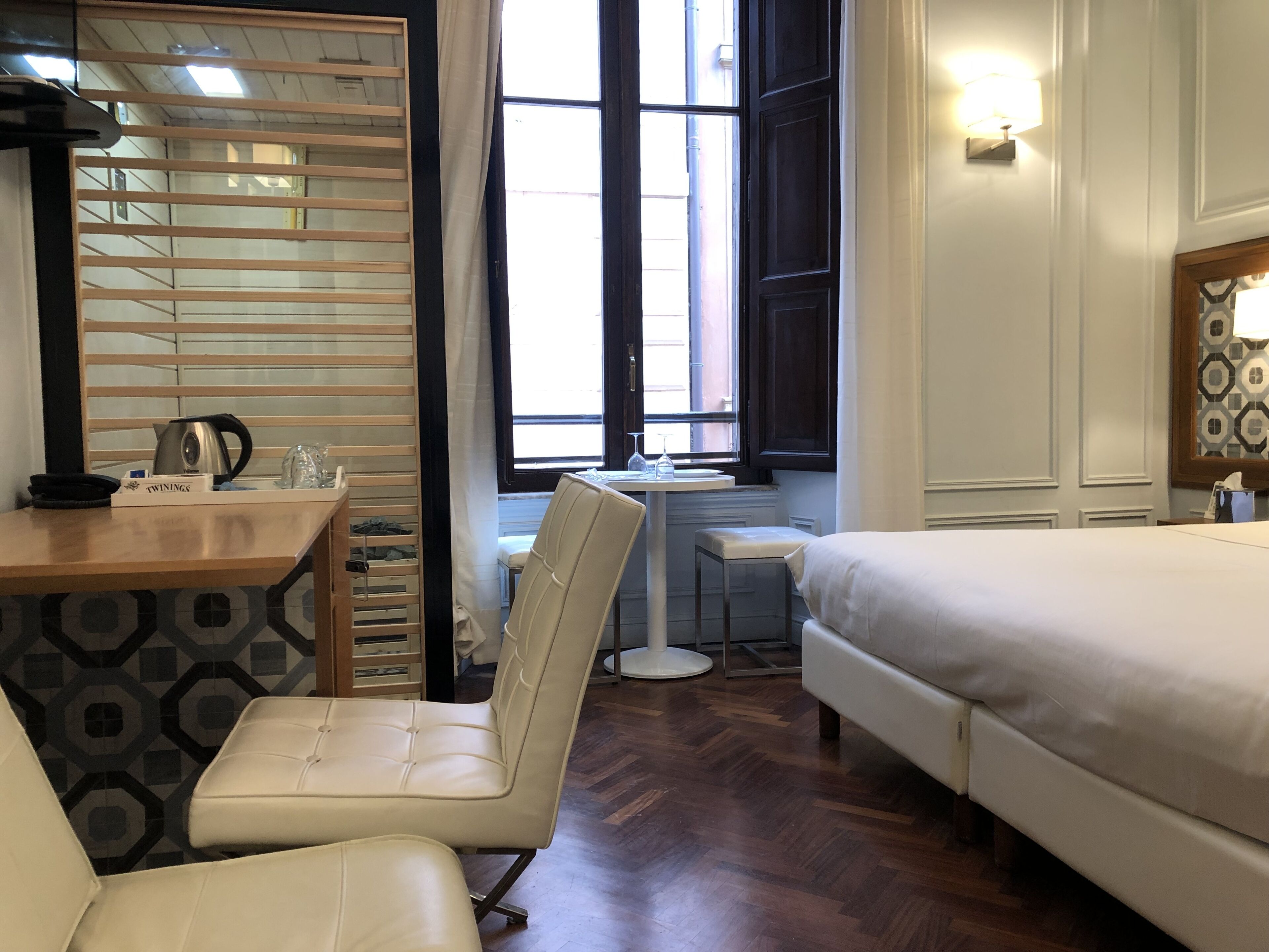 A hotel room with a bed, a desk with a kettle, a chair, a small table with a wine glass, and a window with curtains.