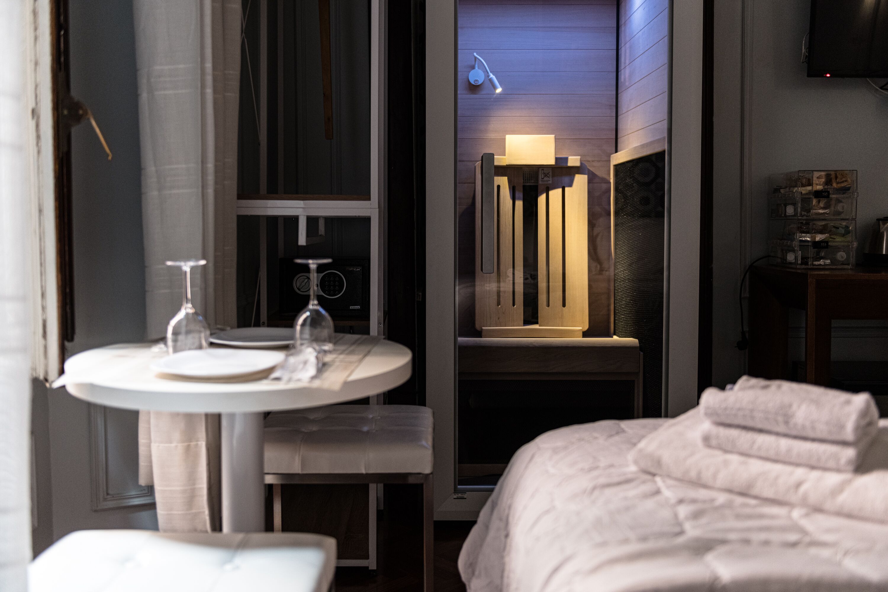 Una camera d'albergo con un letto, un comodino con un bicchiere di vino e un piatto, un tavolino con una sedia, una TV a muro e un armadietto in legno.