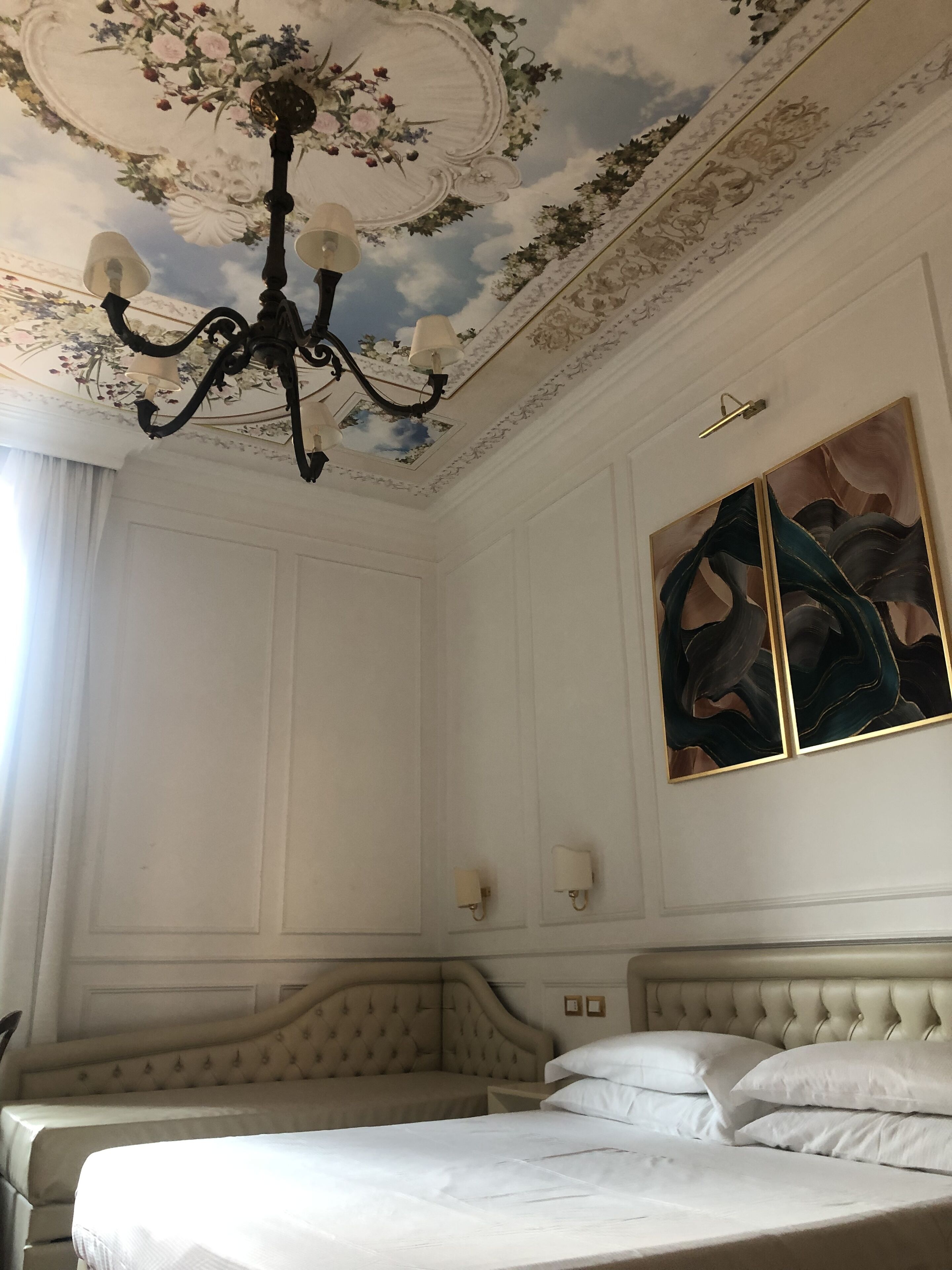 Et soveværelse med en stor seng, en lysekrone, et blomstret loft og et maleri på væggen.