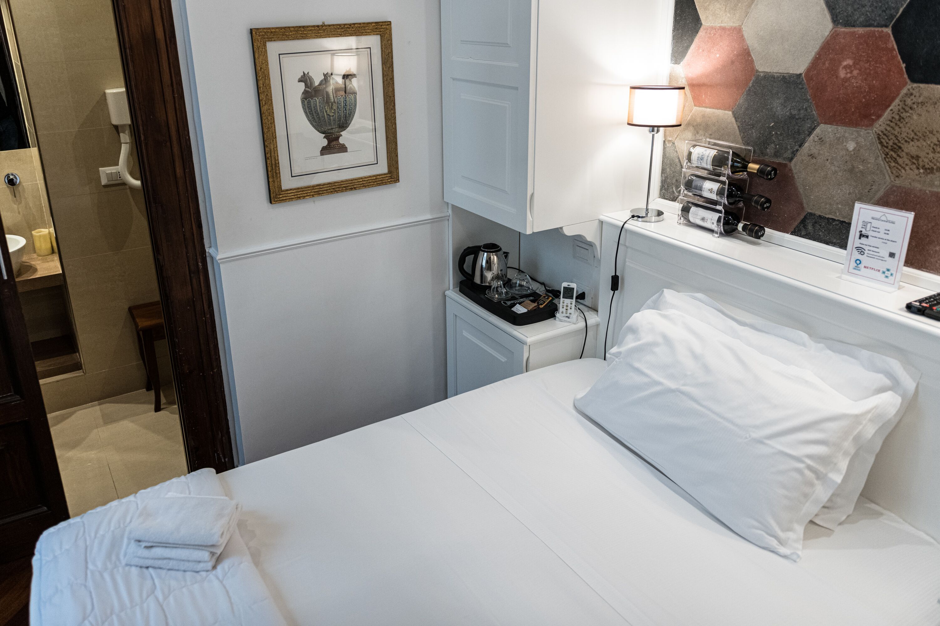 Et hotelværelse med en pænt redt seng, et natbord med lampe, kaffemaskine og vinholder.