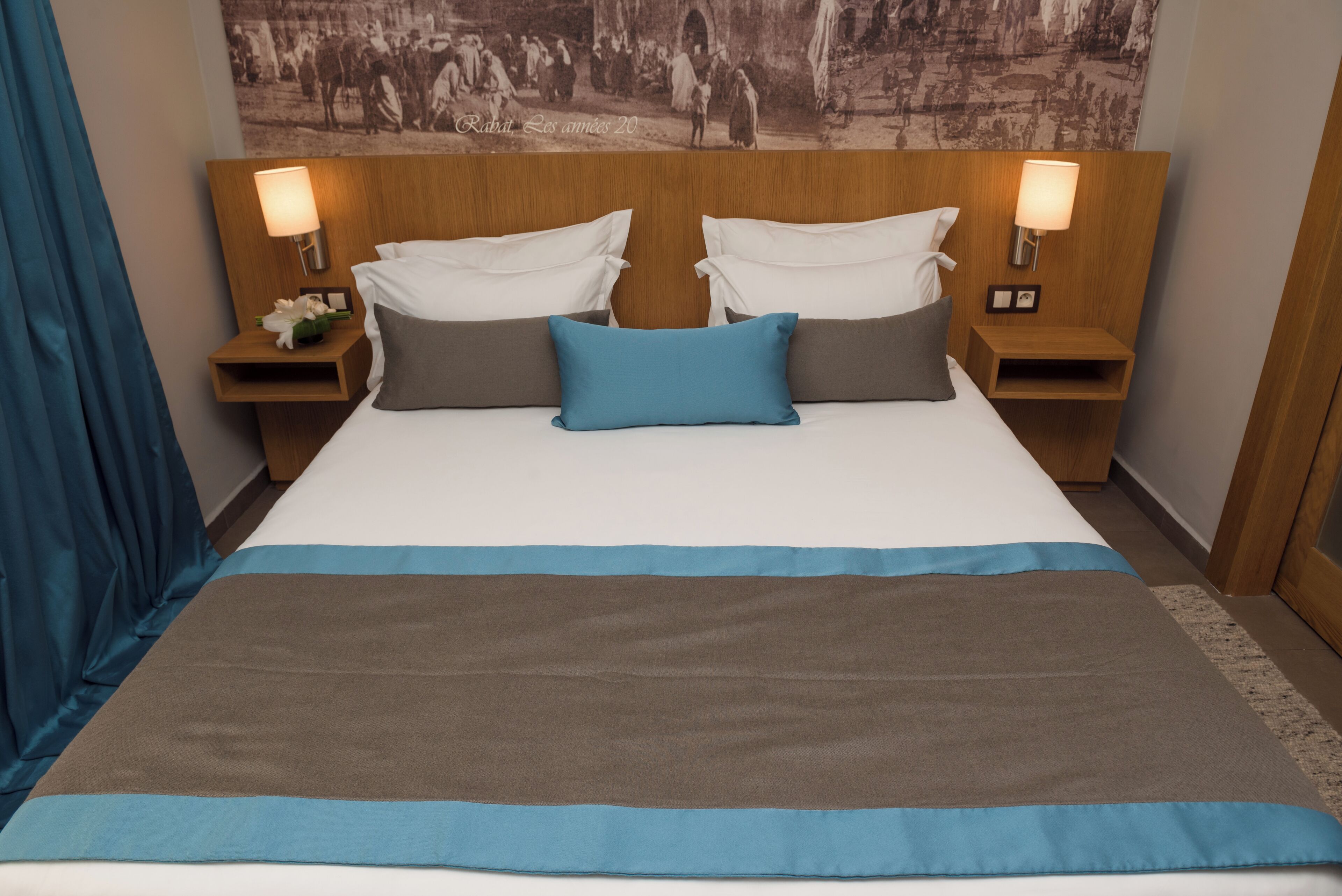 A hotel room with a large bed, two nightstands with lamps, a headboard with a picture, and a blue curtain.