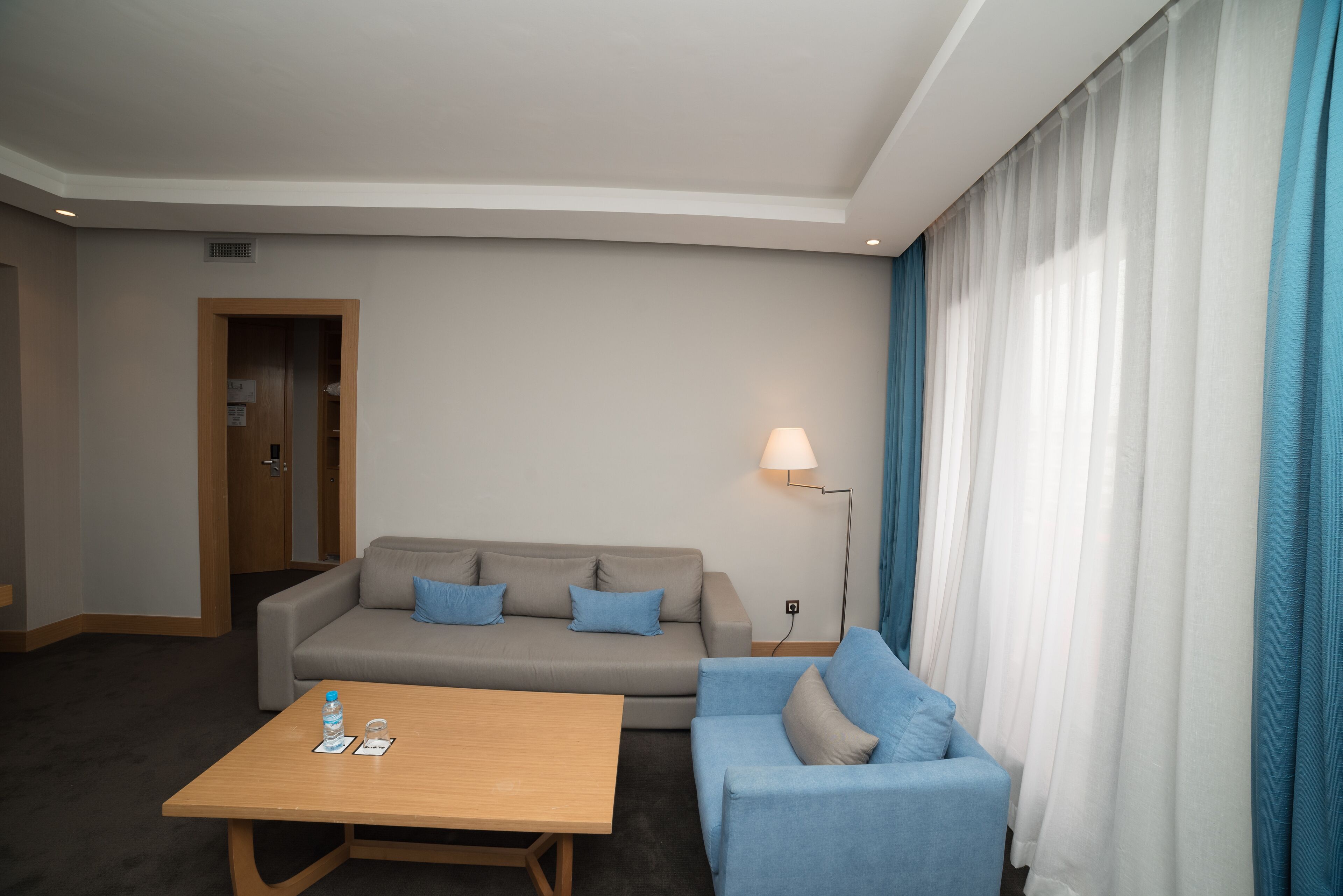 Ein modernes Hotelzimmer mit grauem Sofa, blauem Sessel, Holztisch und Stehlampe.
