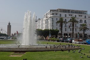 Uma fonte com jatos de água, cercada por um passeio circular. Ao fundo, um edifício branco com varandas e palmeiras.