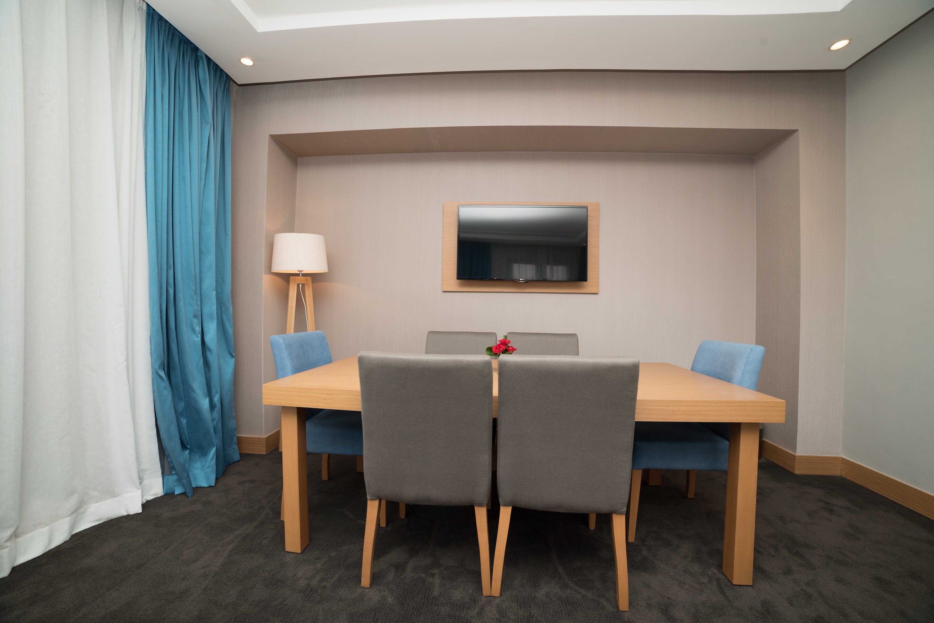 A conference room with a wooden table, grey chairs, a TV mounted on the wall, and a blue curtain.