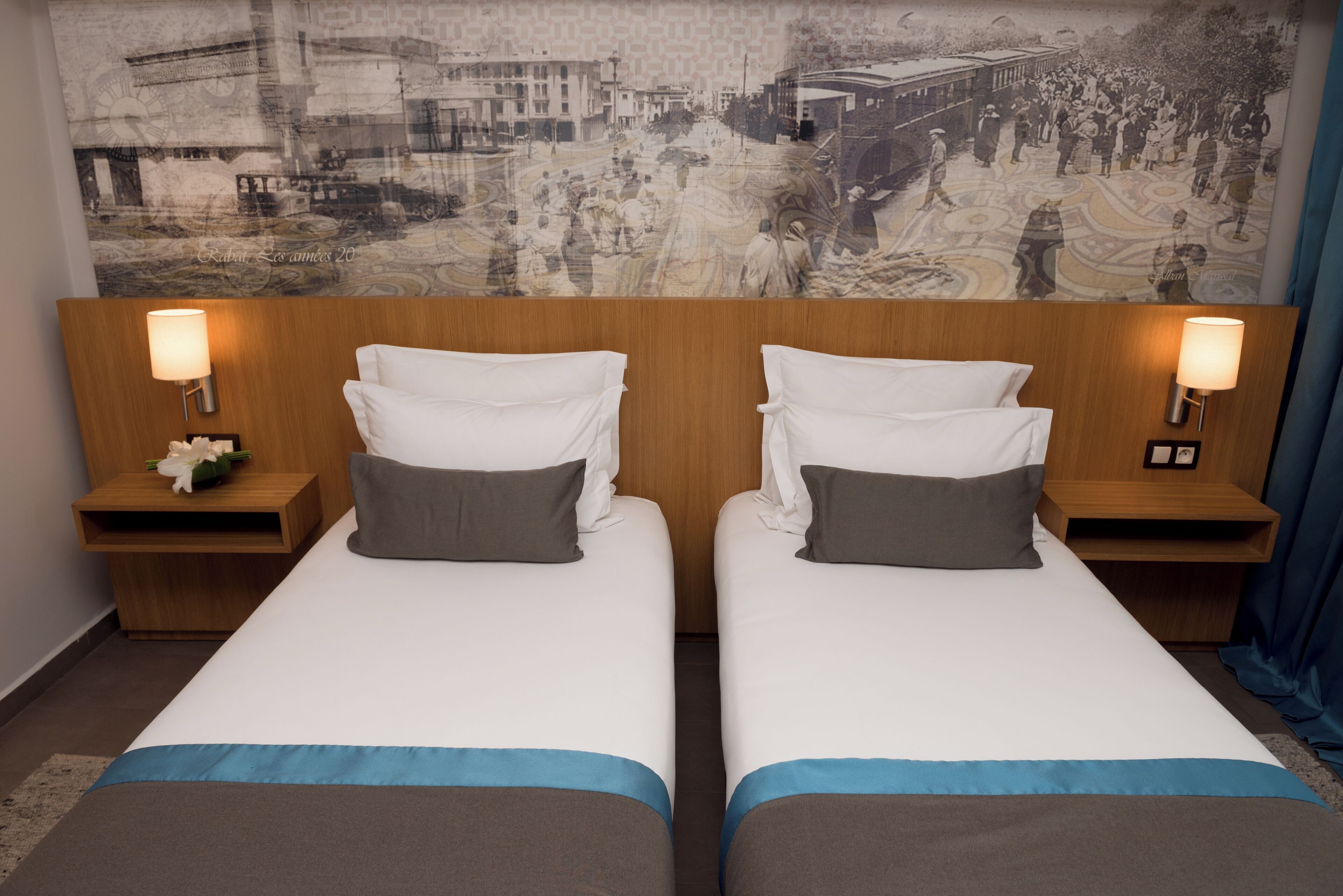 A hotel room with two beds, a headboard with a cityscape mural, bedside lamps, and a nightstand with a flower.