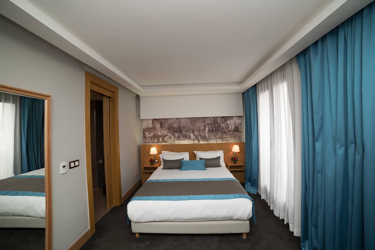 Ein Hotelzimmer mit einem großen Bett, zwei Nachttischen mit Lampen, einer Wandverkleidung am Kopfteil und Blick auf die Stadt durch Vorhänge.