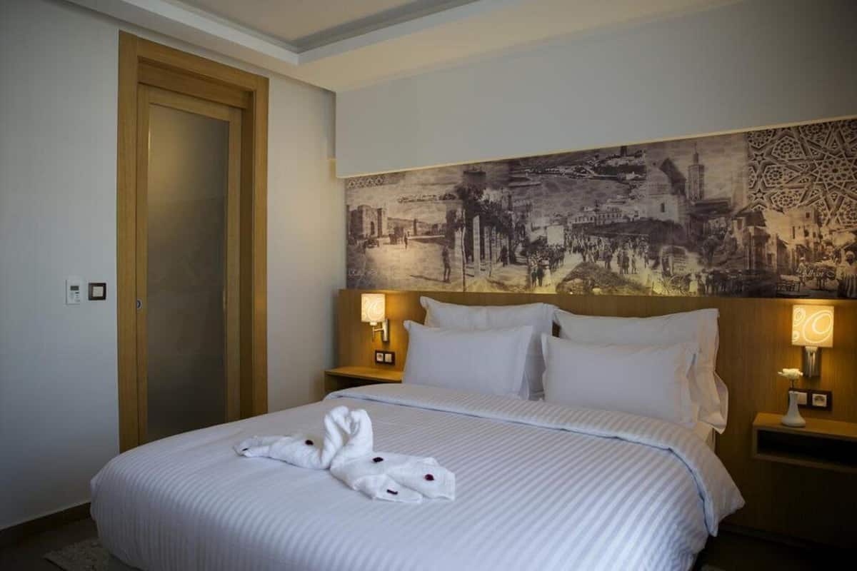 Ein Hotelzimmer mit einem großen Bett, zwei Nachttischlampen, einem Kopfteil mit Stadtansicht als Wandmalerei und einer Holztür.