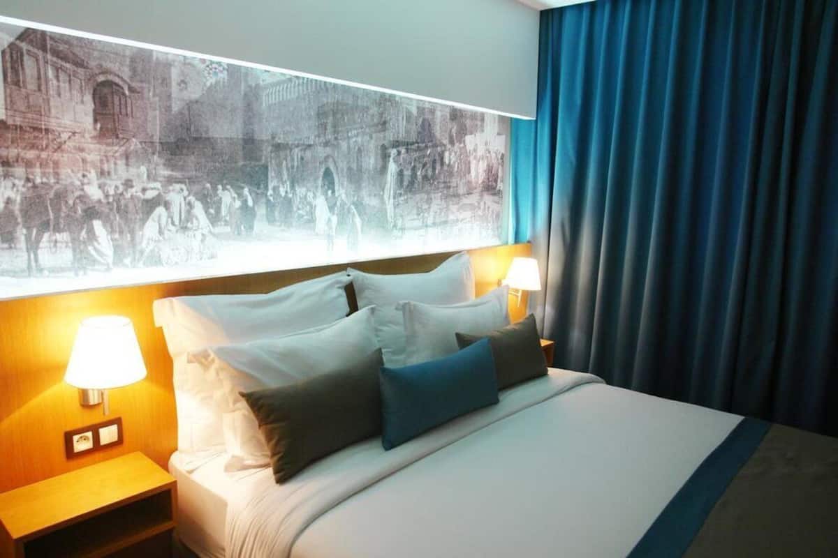 Ein Hotelzimmer mit einem großen Bett, zwei Nachttischlampen, einem Nachttisch und einem großen Fenster mit Blick auf ein historisches Gebäude.