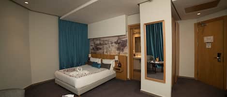 A hotel room with a bed, a desk with a fruit bowl, a chair, a mirror, and a door.
