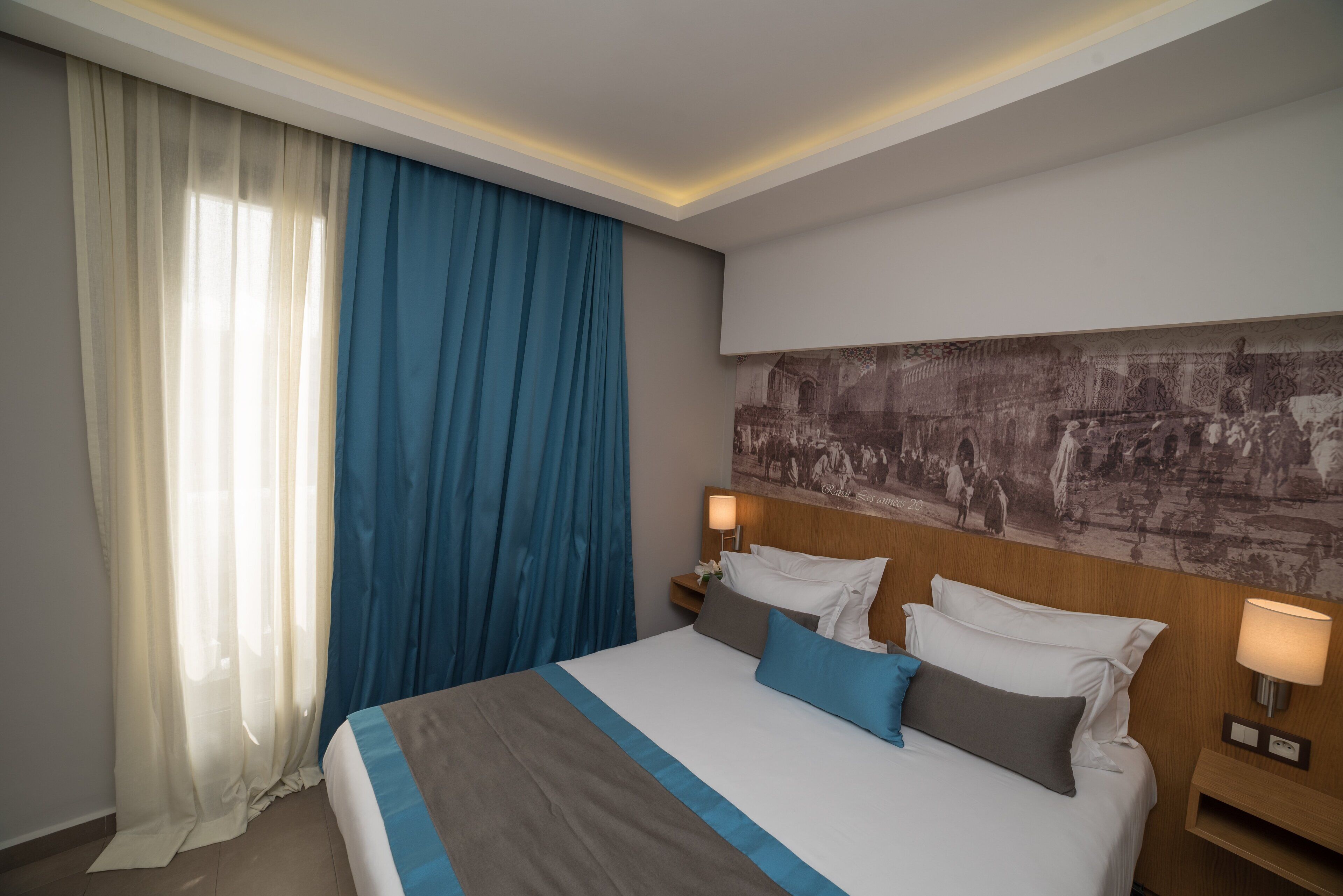 Een hotelkamer met een groot bed, twee nachtkastjes met lampen, een kunstwerk aan de muur en een raam met dunne en blauwe gordijnen.