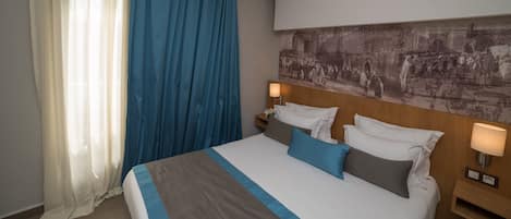 Een hotelkamer met een groot bed, twee nachtkastjes met lampen, een kunstwerk aan de muur en een raam met dunne en blauwe gordijnen.