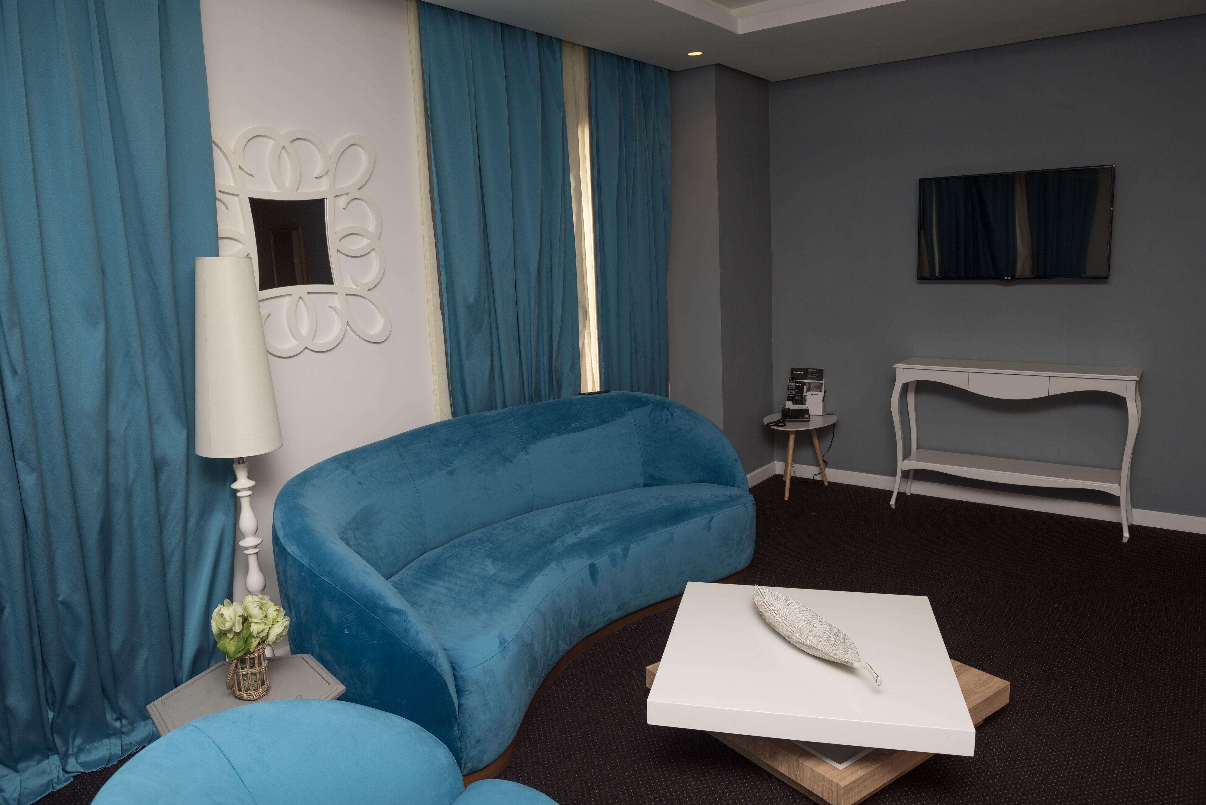 A modern hotel room with a blue sofa, a white coffee table, a lamp, and a TV on the wall.