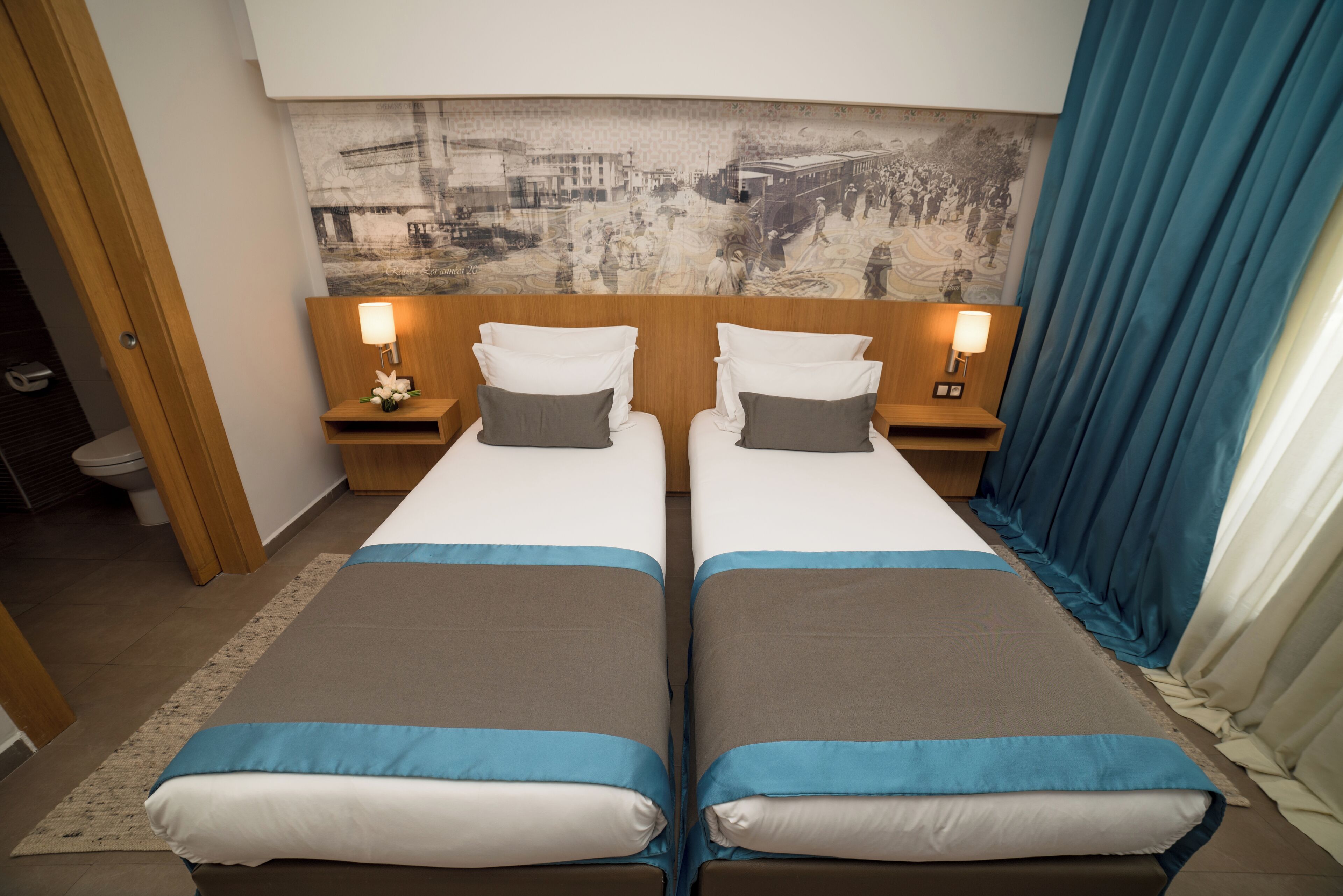 A double room with two beds, nightstands, a wall-mounted lamp, and a large black-and-white mural.