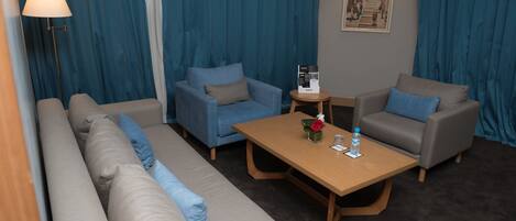 A modern hotel room with a sofa, two armchairs, a small wooden table, a lamp, and a framed picture on the wall.