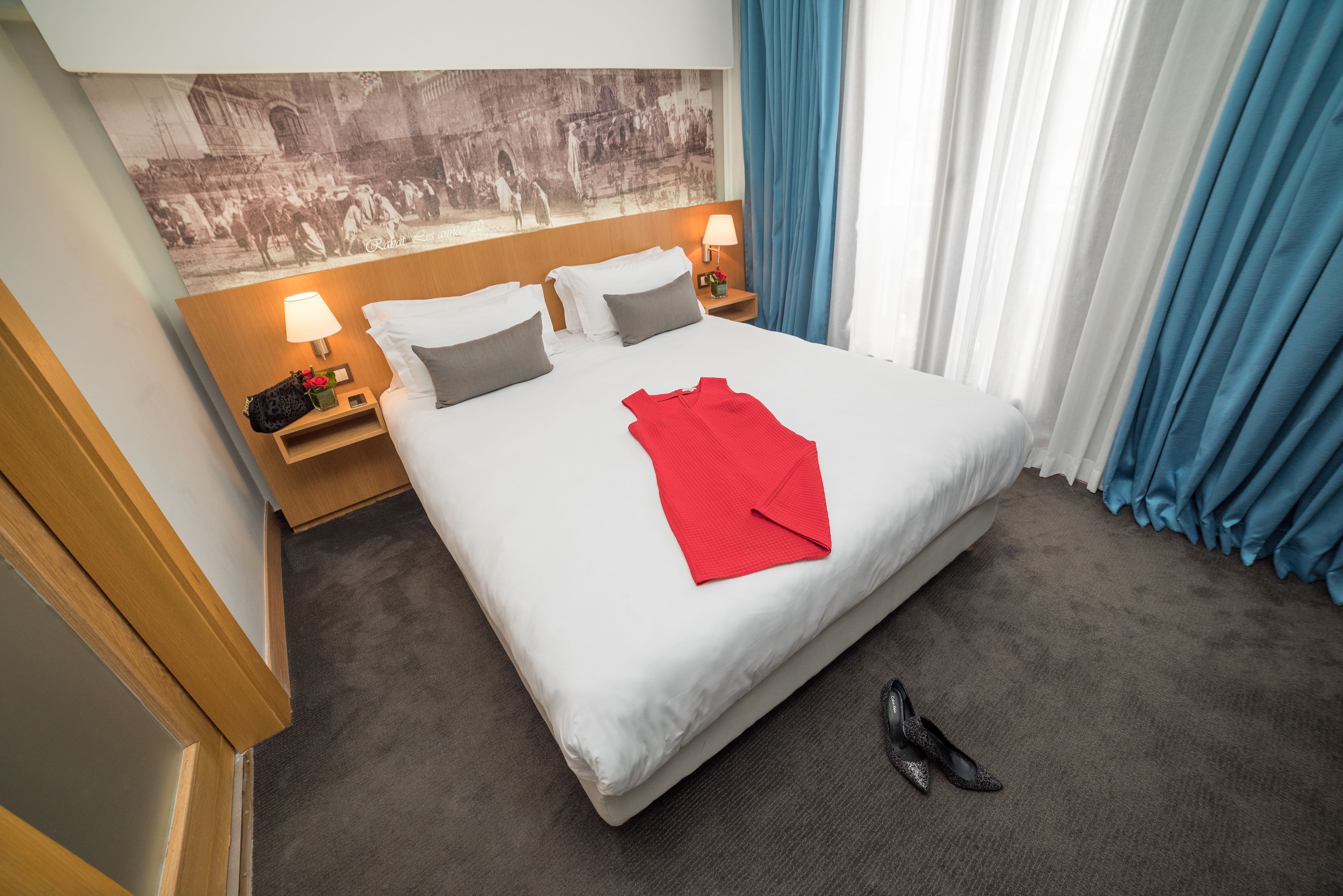Une chambre d’hôtel avec un grand lit, une table de chevet avec une lampe, une serviette rouge et une paire de chaussures posées sur le sol.
