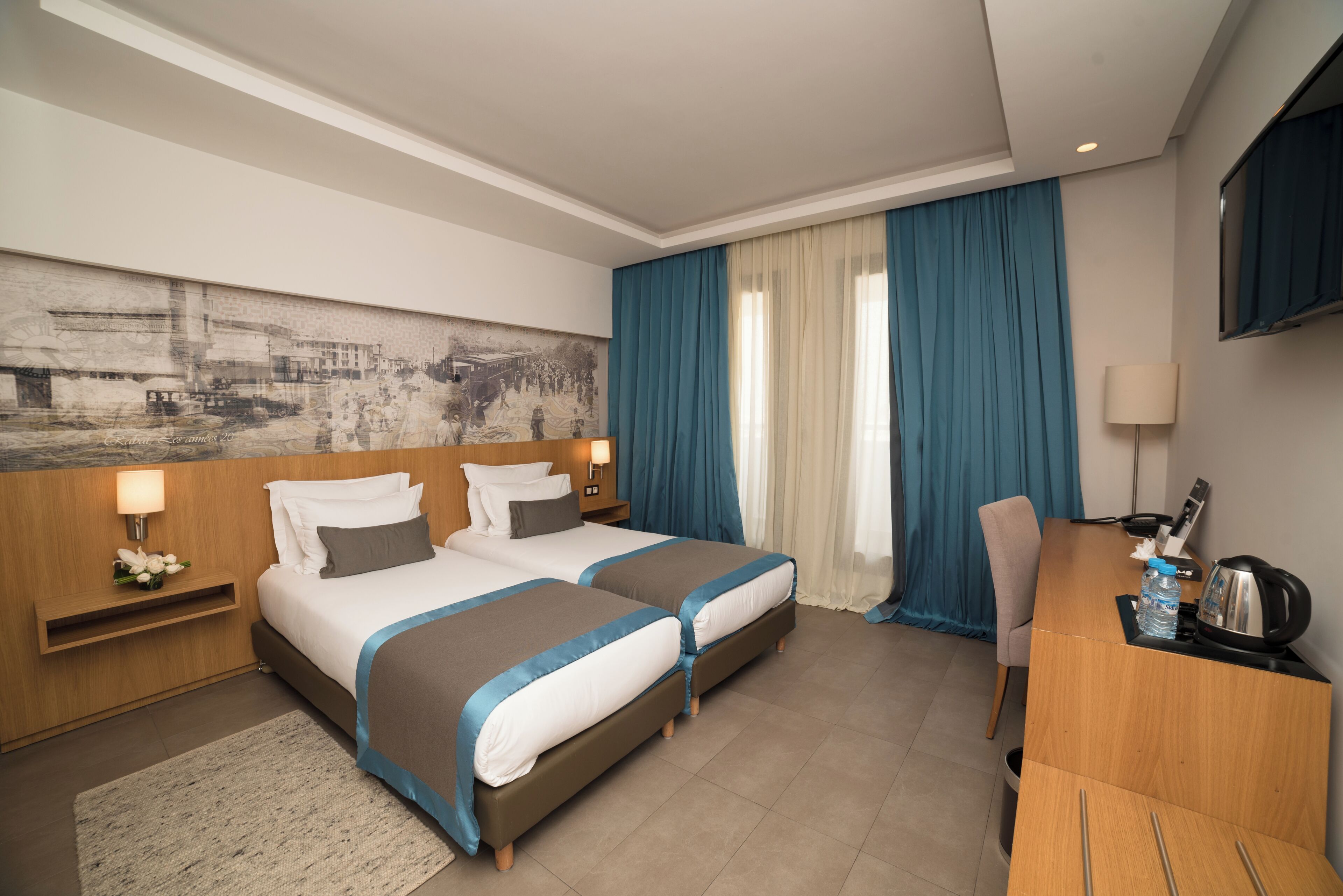 A hotel room with two beds, a desk, a TV, and a large painting on the wall.
