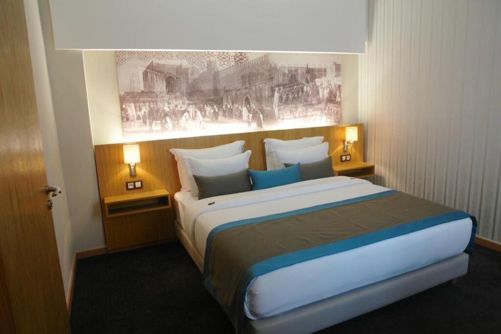 A hotel room with a bed, two bedside tables, a nightstand, a wall-mounted lamp, and a large framed picture above the bed.