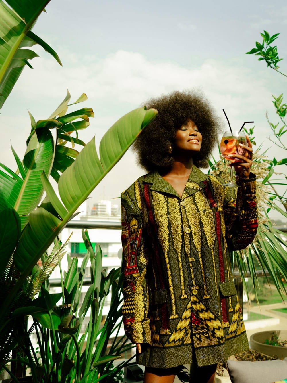 A person standing outdoors with large green plants around, wearing a patterned jacket and holding a drink.