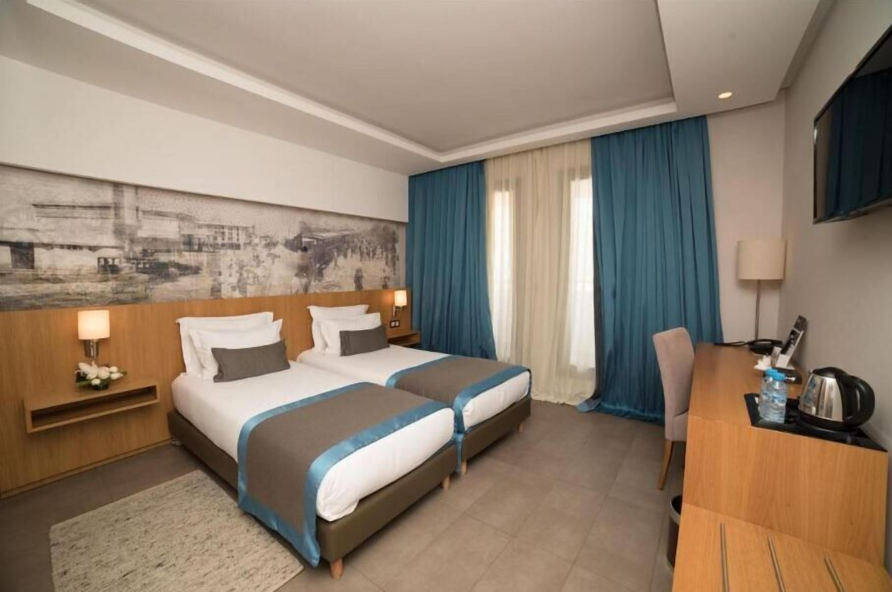 A hotel room with two beds, a desk, a TV, and a painting on the wall.