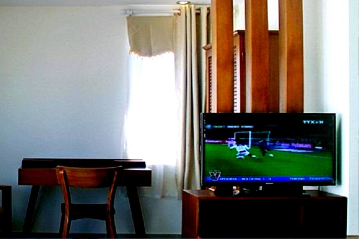A room with a television displaying a sports game, a wooden chair, and a window with curtains.