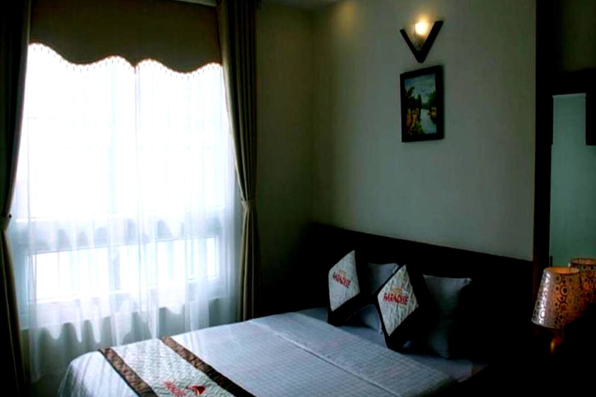 A hotel room with a bed, curtains, a wall-mounted picture, and a nightstand with a lamp.