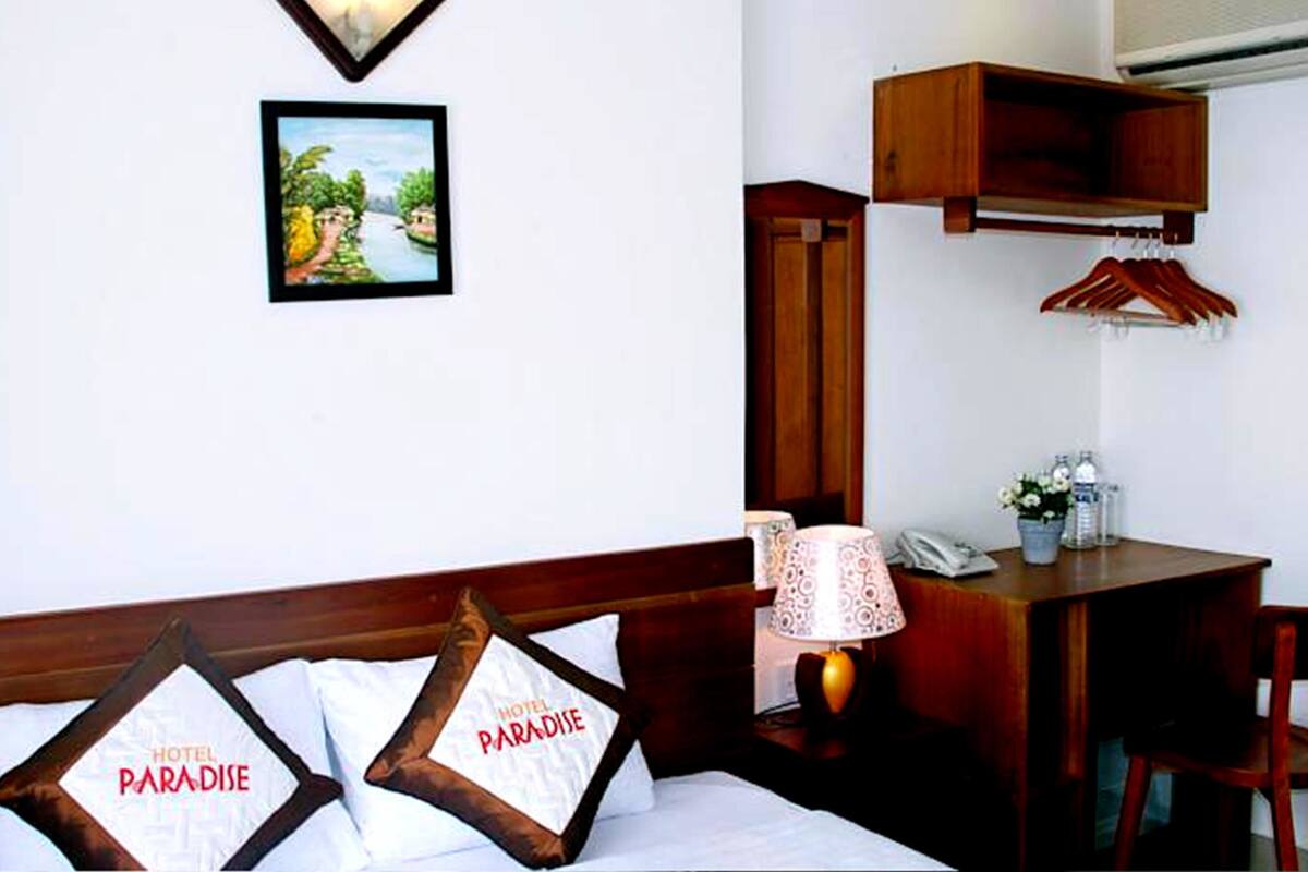 A hotel room with a bed, a desk, a chair, a lamp, a telephone, and a wall-mounted air conditioning unit.