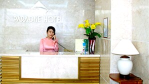 A reception desk at Paradise Hotel with a marble wall, a clock, and a vase with yellow flowers. - Paradise Hotel (Da Nang)