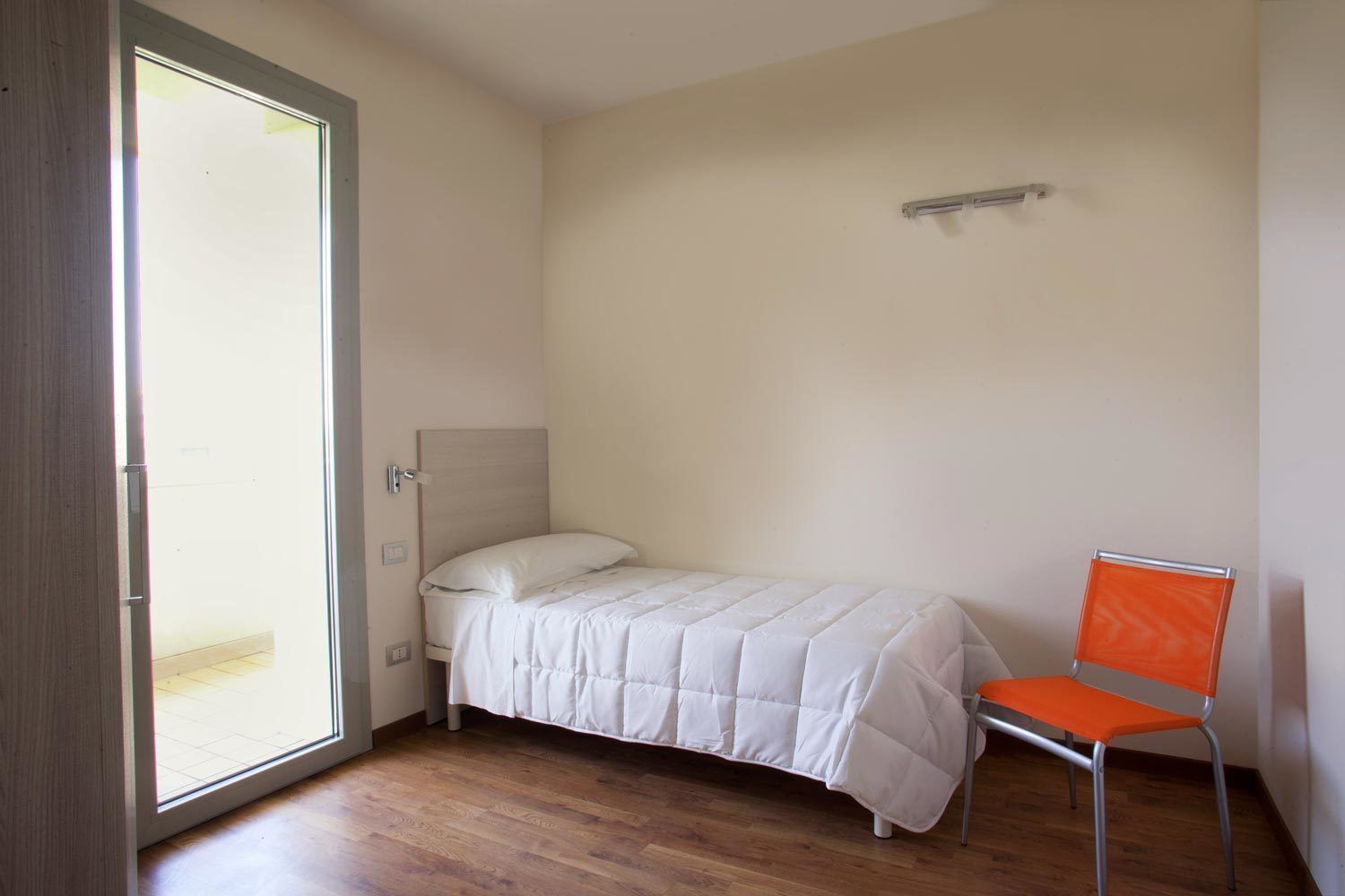 A small, single-bed room with a wooden floor, a white bed, and an orange chair.