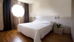 A bedroom with a large bed, wooden flooring, a nightstand with a plant, and a window with curtains. - Residence Le Querce (Cernusco sul Naviglio)