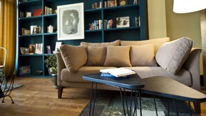 A cozy living room with a beige sofa, a small side table, and a bookshelf filled with books and framed pictures. - Hotel 27 (Tbilisi)