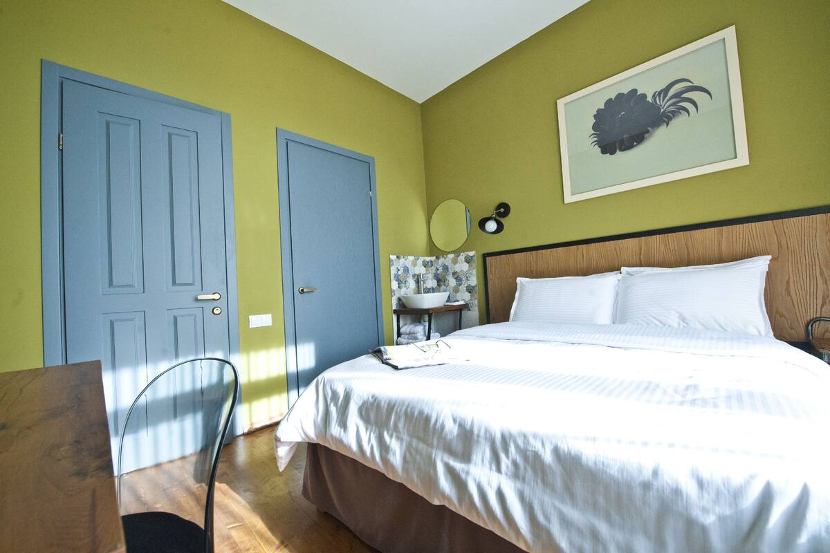 A bedroom with a bed, a chair, a desk, a door, and a wall-mounted mirror.