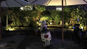 Una scooter estacionada bajo un toldo en un jardín con plantas en macetas y un muro de piedra.