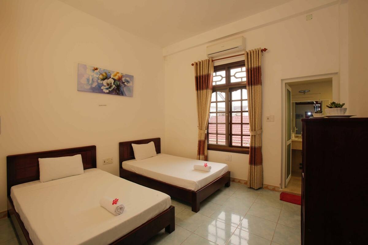 A hotel room with two single beds, a window with curtains, a painting on the wall, and a door leading to a bathroom.