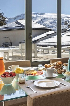 Una mesa de desayuno con diversos alimentos y vistas a montañas nevadas a través de amplios ventanales.