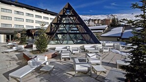 Un edificio moderno con fachada de vidrio triangular, rodeado de terrazas exteriores y montañas nevadas al fondo.