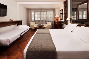 Habitación de hotel con una cama grande, una cama más pequeña, un escritorio, una silla, un televisor y una ventana con cortinas.