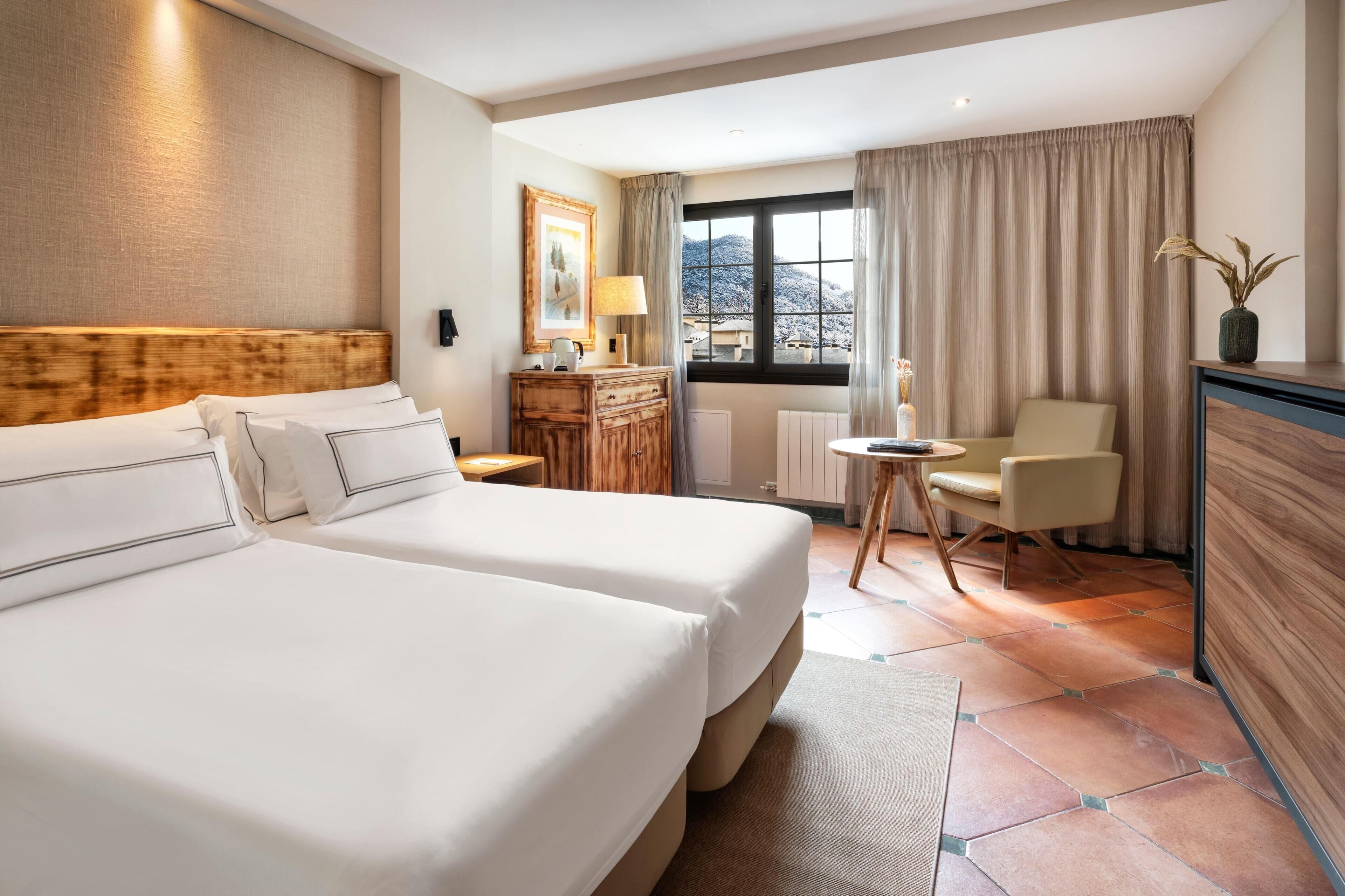 Habitación de hotel con dos camas, cabecera de madera, escritorio con silla, ventana con cortinas y vistas al exterior.