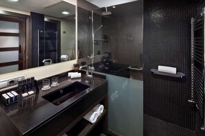 Un baño moderno con ducha revestida de azulejos oscuros, un espejo grande, un lavabo con artículos de aseo y un perchero.