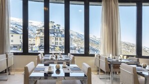 Un restaurante con mesas y sillas dispuestas para comer, ventanas amplias con vistas a montañas nevadas y edificios.