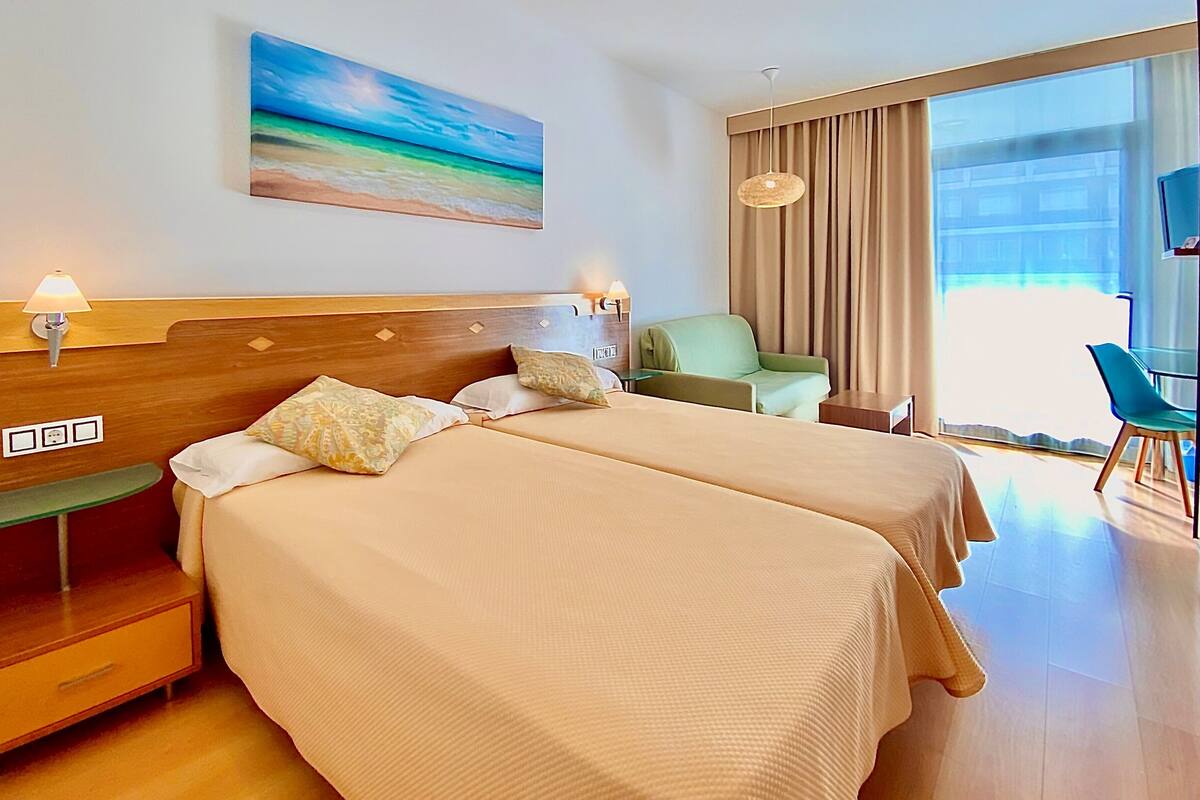 A hotel room with two beds, a green armchair, a TV, and a large window.