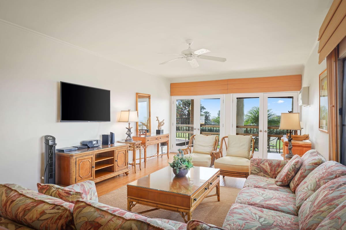 A spacious living room with a flat-screen TV, wooden furniture, a ceiling fan, and large windows offering a view of the outdoors.
