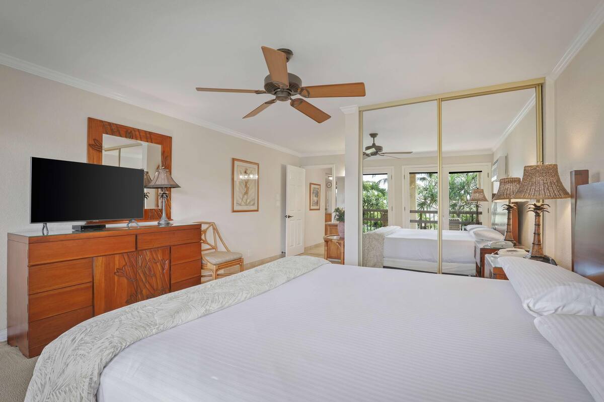 A hotel room with a large bed, a TV on a wooden dresser, a ceiling fan, and a large mirror reflecting the room.