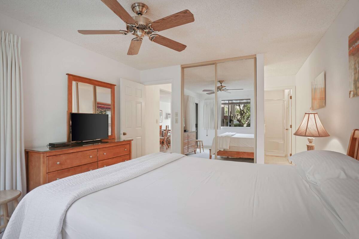 A hotel room with a bed, a television on a wooden dresser, a ceiling fan, and a large mirror.