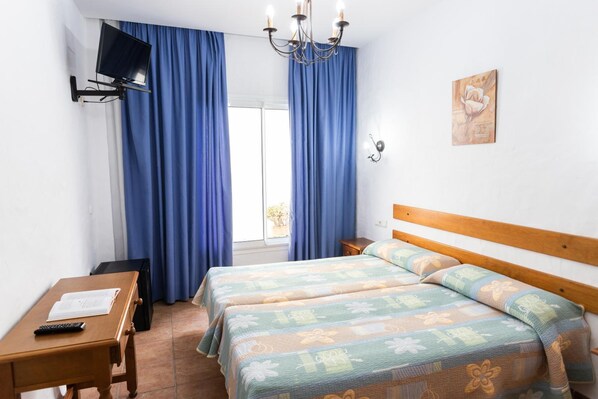 A hotel room with two beds, a TV mounted on the wall, a desk with a remote control, and a window with blue curtains. - Oasis Atalaya (Conil de la Frontera)