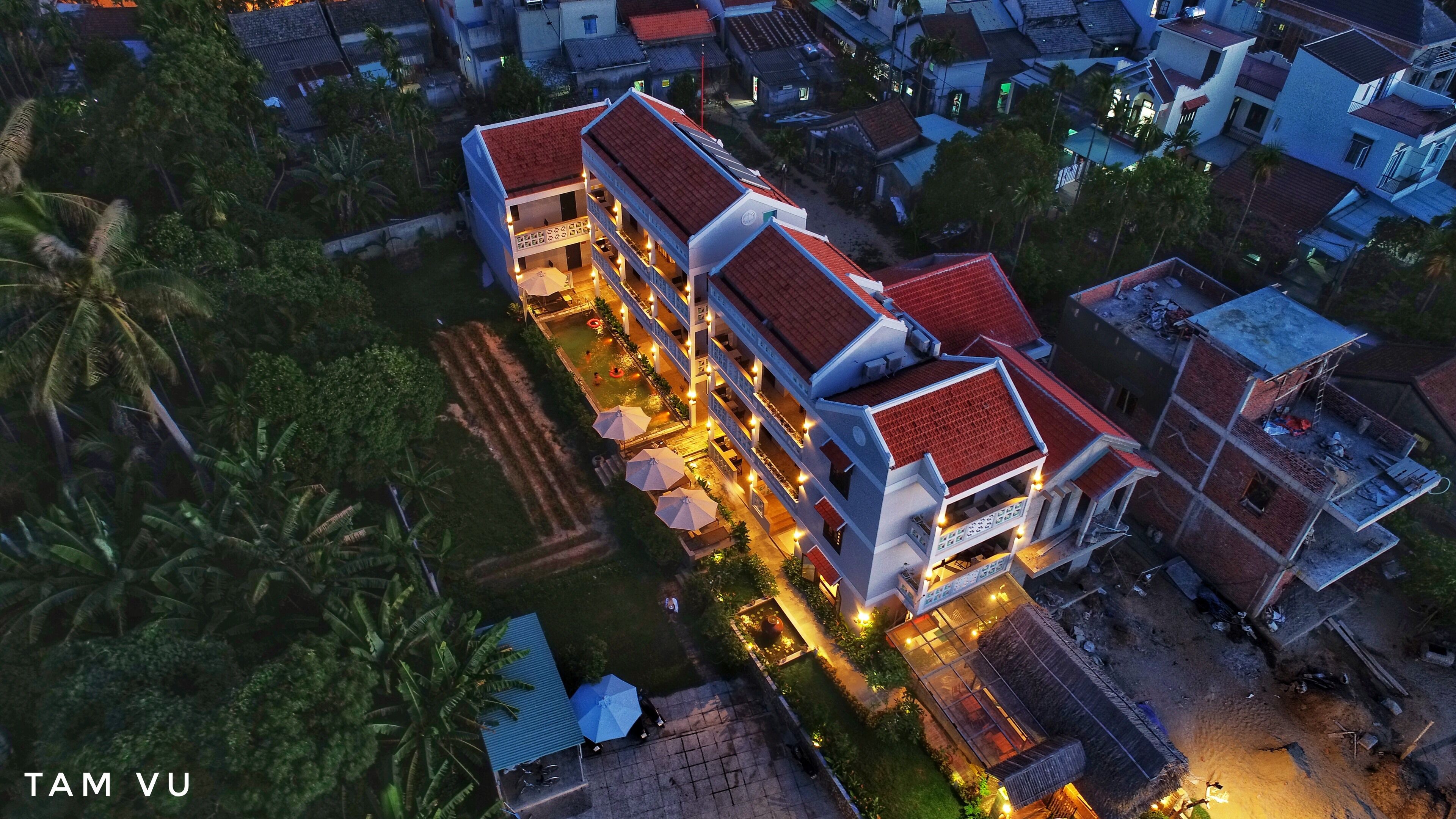 An aerial view of a multi-story hotel with a red-tiled roof, surrounded by greenery and other buildings.