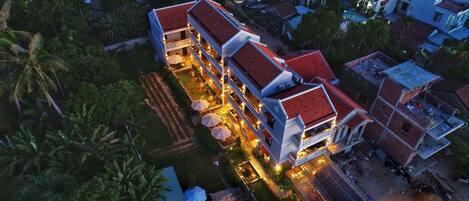 An aerial view of a multi-story hotel with a red-tiled roof, surrounded by greenery and other buildings.