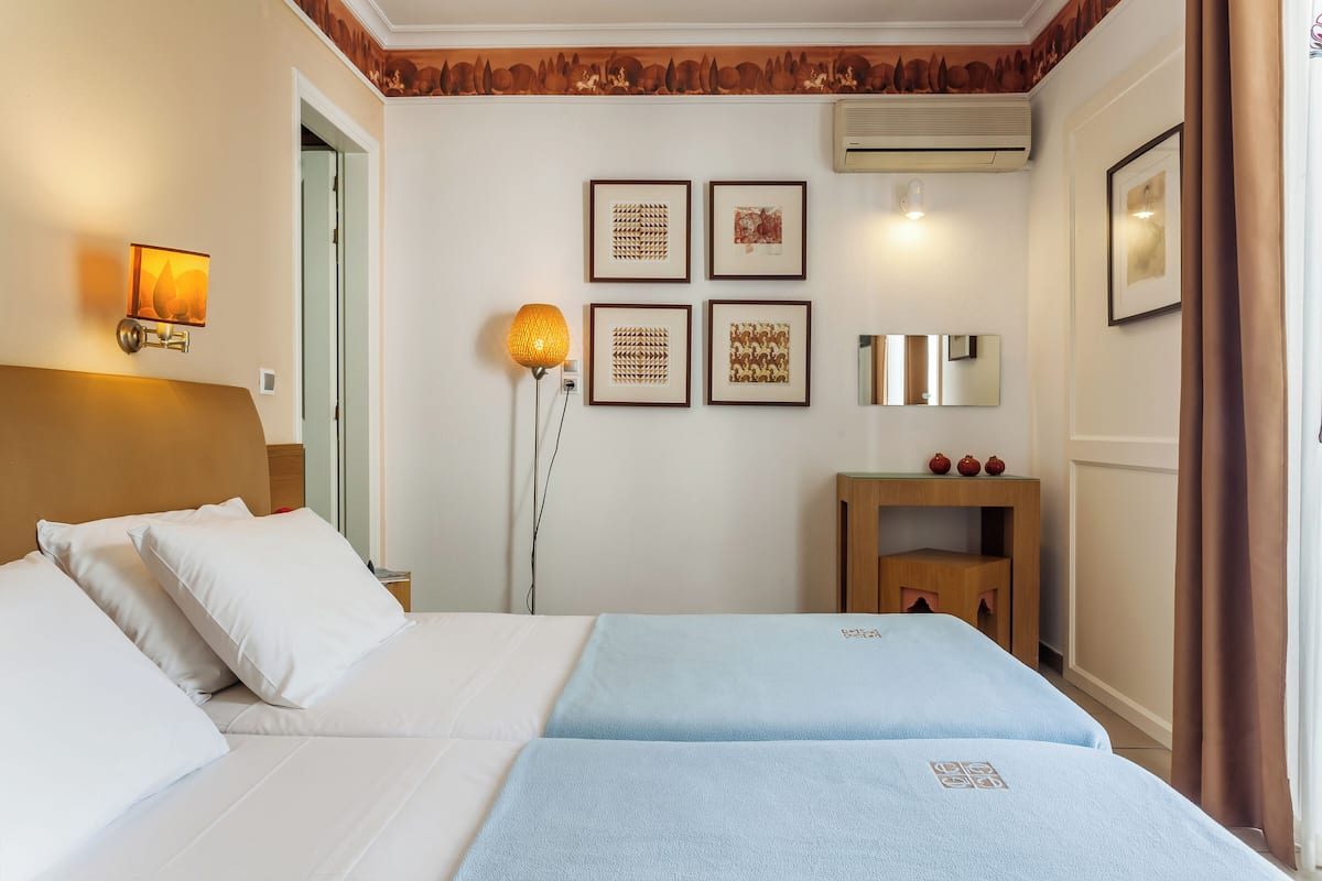 A hotel room with two beds, a nightstand, a lamp, framed artwork on the wall, and a small wooden table with decorative items.